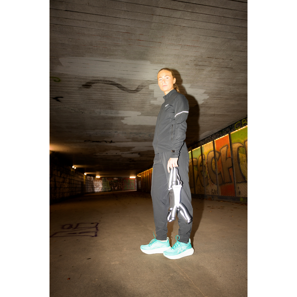 En person i sort træningsdragt og turkise sneakers står i en graffitibelagt tunnel med løbesko og BeeSafe LED Harness USB Phone Pocket (Unisex, Silver) på, klar til natlige løbeture under ovenlys.