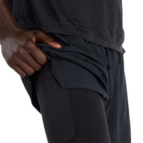 En person iført New Balance Rc Lined Sh 7In løbeshorts til herre i sort løfter det yderste lag for at afsløre et tætsiddende indre lag, med et nærbillede af deres hånd og overkrop mod en almindelig, ryddelig baggrund.