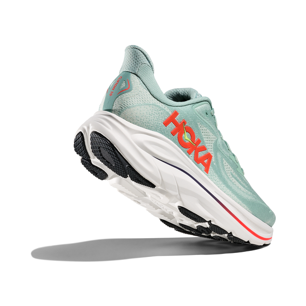 En Hoka Clifton 10, Herre i Sage/Neon Flame med en tyk hvid sål, sort slidbane og fed orange branding vises fra en vinkel bagfra mod en almindelig hvid baggrund.