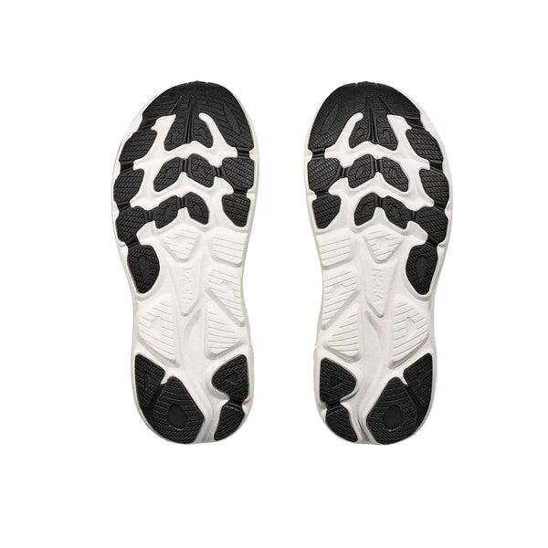 Et par Hoka Clifton 10 Dame Black/White løbesko er vist med sålerne vendt opad. De hvide såler har et sort, uregelmæssigt segmenteret mønster, der giver greb og trækkraft. Billedet er taget på en hvid baggrund.