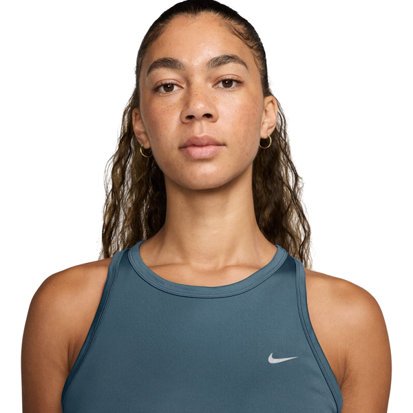 En kvinde med bølget hår er iført en Nike Dri-Fit Running Tank Top for dames i Smokey Blue/Reflective Silver med fugtstyring og åndbarhed. Hun har også guldøreringe og ser neutralt ud mod en almindelig hvid baggrund.