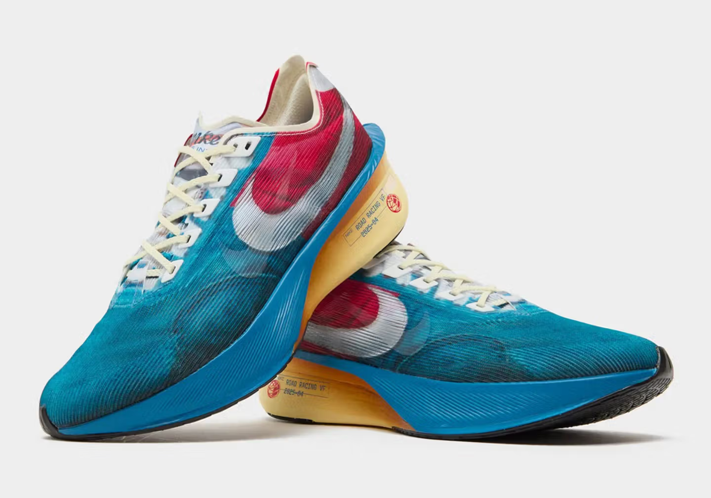Et par Nike-løbesko med blå mesh-overdel, fremtrædende hvide swooshes, røde detaljer nær hælen, cremefarvede såler og hvide snørebånd. Den ene sko hviler oven på den anden og viser detaljer på siden og sålen.