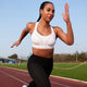 En kvinde i sportstøj løber på en udendørs bane iført Shock Absorber Ultimate Run Bra Padded, Dame, White for maksimal støtte sammen med sorte leggings. Scenen omfatter græs, træer og en klar himmel i baggrunden.