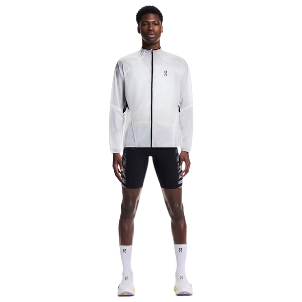 En mand står og kigger fremad iført On Zero Jacket, Herre, White med sorte sportsshorts, hvide strømper og lyse sneakers på en almindelig hvid baggrund.