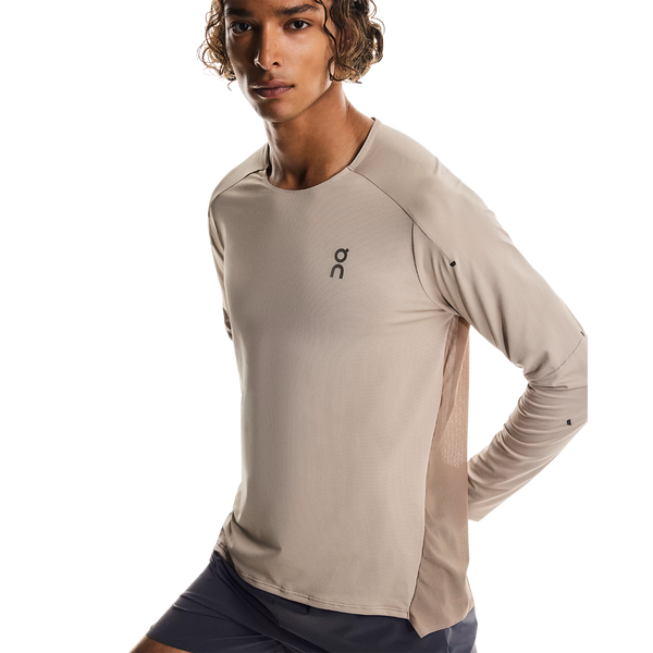 En person med krøllet hår bærer On Performance Long-T, Herre, Desert/Cinder med mørke shorts på en hvid baggrund - det hurtigtørrende materiale gør den perfekt til træning i køligere temperaturer.