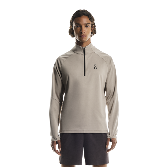 En person ser frem for sig iført en On Climate Shirt, Herre, Desert - en beige langærmet top med kvart lynlås og et lille logo på venstre bryst - sammen med mørke shorts på en almindelig hvid baggrund.