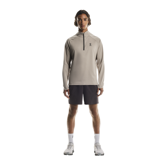 En person står med ansigtet fremad i en On Climate Shirt, Herre, Desert, sammen med mørke shorts, hvide strømper og hvide sportssko på en almindelig hvid baggrund.