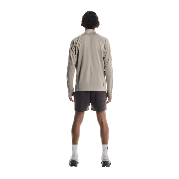 En person står med ryggen til, iført On Climate Shirt, Herre, Desert (lysegrå zip-up), med mørke shorts, hvide sokker og sportssko på en almindelig hvid baggrund.