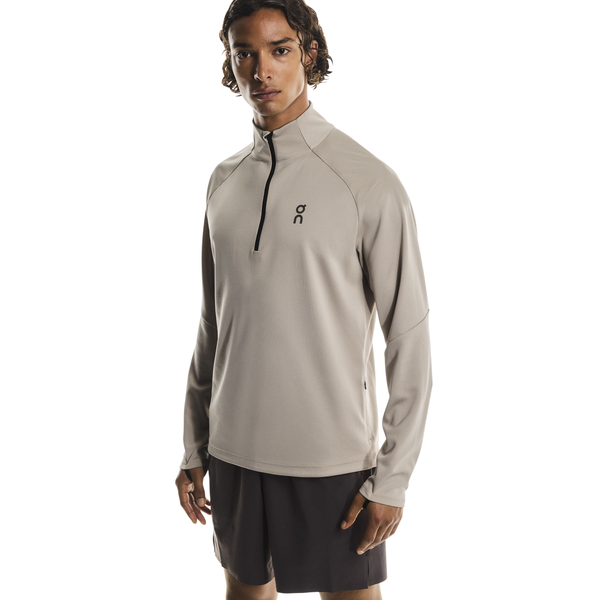 En person iført On Climate Shirt, Herre, Desert - en lys beige top med et lille sort logo på venstre bryst - sammen med sorte shorts og stående på en almindelig hvid baggrund.