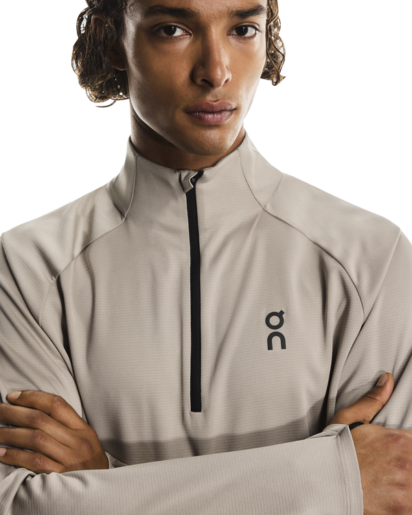 En person med krøllet hår og korslagte arme bærer On Climate Shirt Herre i Desert med sort lynlås og brystlogo og står på en hvid baggrund.