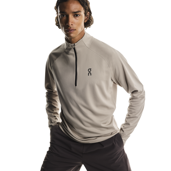 En person står på en hvid baggrund iført On Climate Shirt, Herre, Desert - en beige langærmet sportstop med kvart lynlås og sort logo på brystet - sammen med mørke bukser og hænderne hvilende på hofterne.