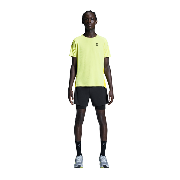 En person ser frem for sig i en lysegul sportstee, On 5" Performance 2/1 Shorts til mænd i sort (åndbart materiale), sorte sokker og grå løbesko. Baggrunden er hvid og ren.