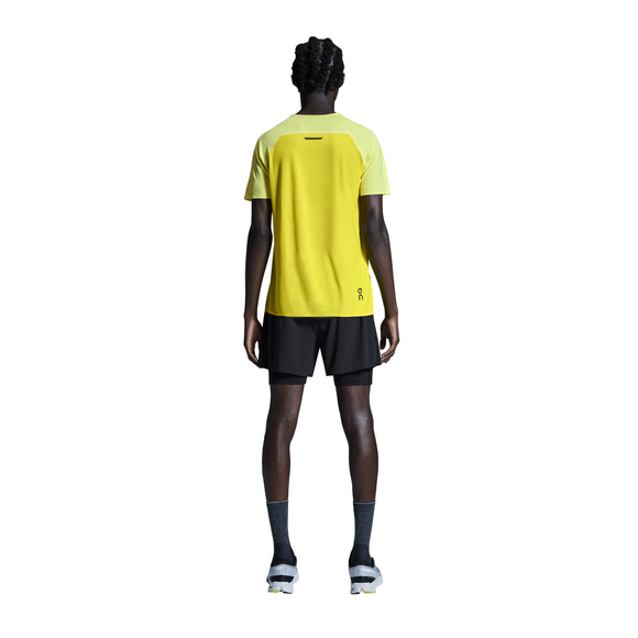 En person står med ansigtet vendt mod kameraet iført en gul kortærmet skjorte, On 5" Performance 2/1 Shorts Herre i sort, grå sokker og hvide sneakers på en hvid baggrund.