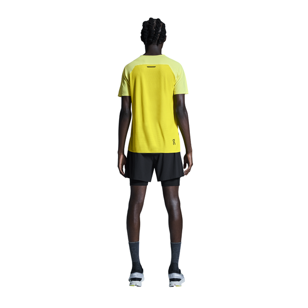 En person står med ansigtet vendt mod kameraet iført en gul kortærmet skjorte, On 5" Performance 2/1 Shorts Herre i sort, grå sokker og hvide sneakers på en hvid baggrund.