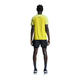 En person står med ansigtet vendt mod kameraet iført en gul kortærmet skjorte, On 5" Performance 2/1 Shorts Herre i sort, grå sokker og hvide sneakers på en hvid baggrund.