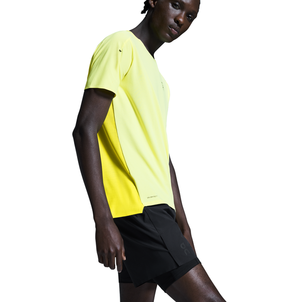 En person iført en gul sports-T-shirt og On 5" Performance 2/1 Shorts, Herre, Black står på en hvid baggrund og kigger let nedad med den ene arm afslappet langs siden.