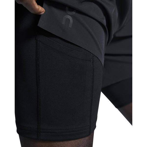 Nærbillede af en person i On 5" Performance 2/1 Shorts, Herre, Black, der løfter det yderste lag for at vise en lomme på indersiden. Løbeshortsene har synlige syninger, glat tekstur og åndbart materiale for øget komfort.