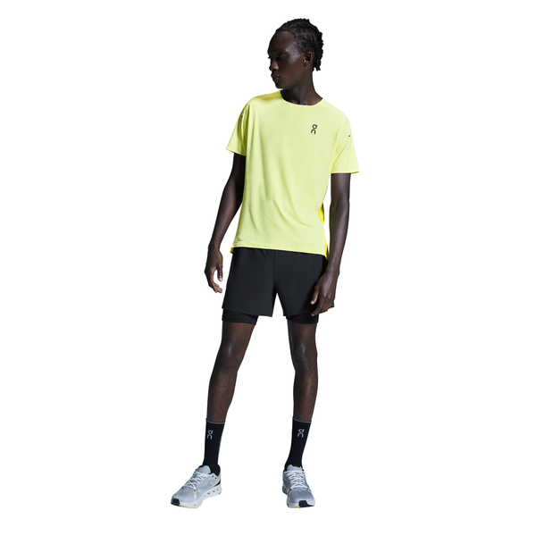 En person iført en gul atletisk t-shirt, On 5" Performance 2/1 Shorts Herre Black, sorte sokker og grå sneakers står med ansigtet lidt til siden mod en hvid baggrund.