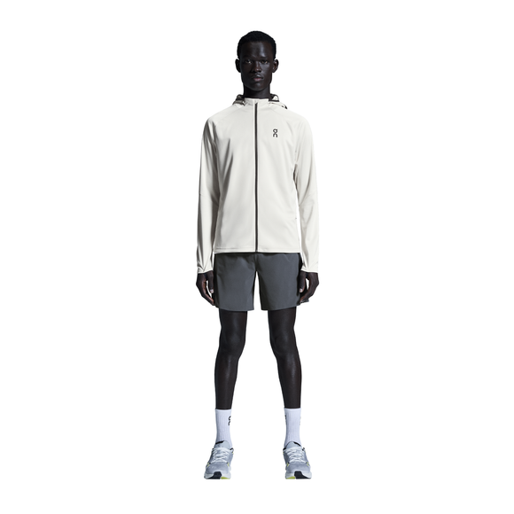 En person står med ansigtet fremad iført en lys jakke med hætte, On 7" Performance Shorts til mænd i Eclipse, hvide strømper og lysegrå sportssko på en almindelig hvid baggrund.
