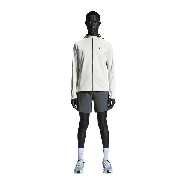 En person står med ansigtet fremad iført en lys jakke med hætte, On 7" Performance Shorts til mænd i Eclipse, hvide strømper og lysegrå sportssko på en almindelig hvid baggrund.
