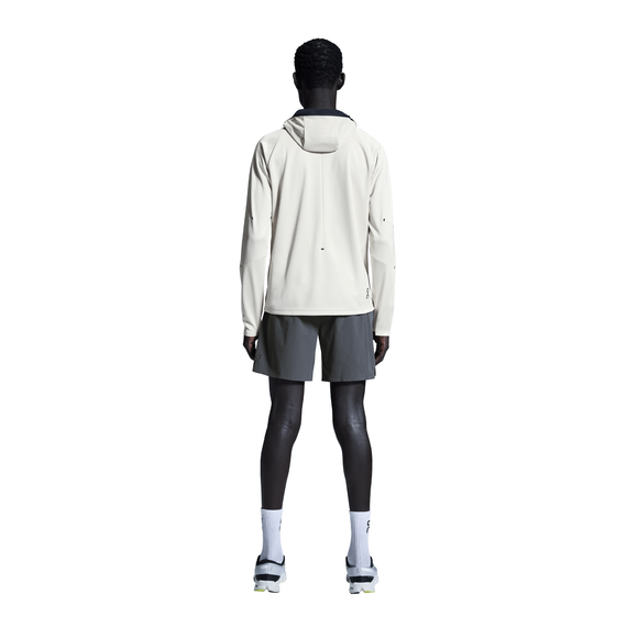 En person står med ansigtet vendt mod kameraet iført en lys jakke med hætte, On 7" Performance Shorts til mænd i Eclipse, hvide sokker og sportssko på en almindelig hvid baggrund.