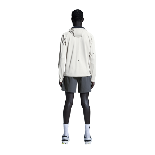 En person står med ansigtet vendt mod kameraet iført en lys jakke med hætte, On 7" Performance Shorts til mænd i Eclipse, hvide sokker og sportssko på en almindelig hvid baggrund.