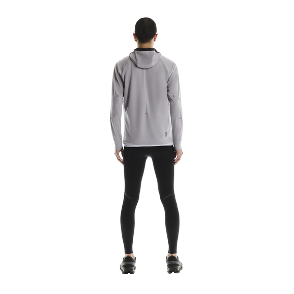 En person står med ryggen til kameraet iført en lysegrå jakke med hætte, On Performance Tights Herre i sort og sorte sneakers på en almindelig hvid baggrund.