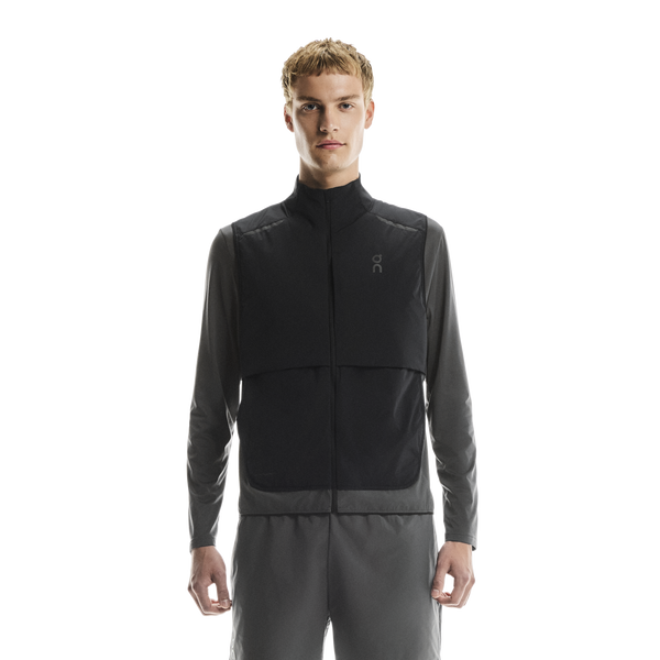 En mand står med front mod en almindelig hvid baggrund iført On Weather Vest, Herre, Black/Eclipse over en langærmet grå skjorte og matchende grå bukser.