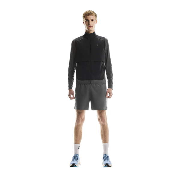 En person kigger fremad og er iført On Weather Vest, Herre, Black/Eclipse, grå sportsshorts, hvide strømper og grå og blå løbesko på en almindelig hvid baggrund.