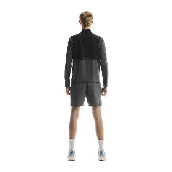 En person med kort blondt hår ser væk fra kameraet og er iført On Weather Vest, Herre, Black/Eclipse, sammen med sorte shorts, hvide sokker og hvide sportssko på en almindelig hvid baggrund.