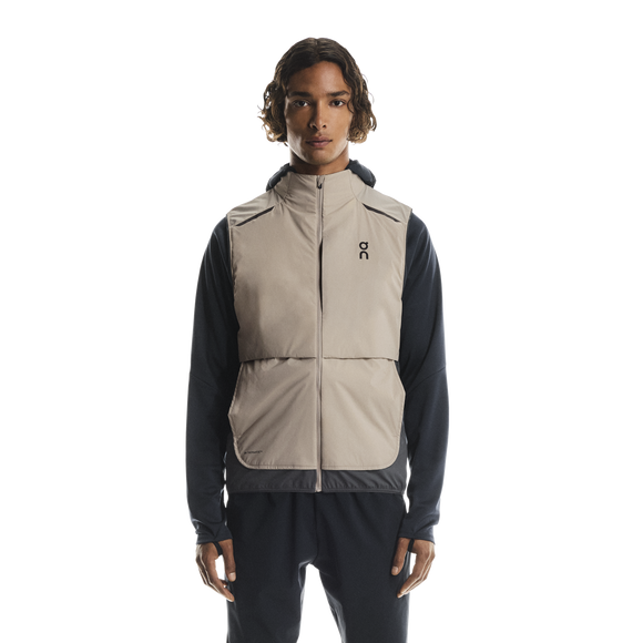 En mand ser frem for sig iført On Weather Vest, Herre, Desert/Eclipse over en langærmet sort skjorte og sorte bukser på en almindelig hvid baggrund.