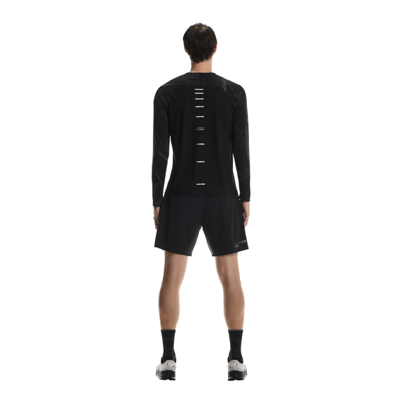 En person kigger væk fra kameraet iført On Performance Long-T Lumos, Herre, Black, med vandrette hvide striber ned ad ryggen, sammen med sorte shorts, sorte sokker og hvide sportssko på en almindelig hvid baggrund.