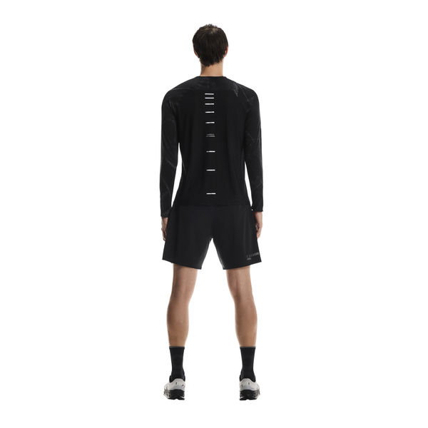 En person kigger væk fra kameraet iført On Performance Long-T Lumos, Herre, Black, med vandrette hvide striber ned ad ryggen, sammen med sorte shorts, sorte sokker og hvide sportssko på en almindelig hvid baggrund.
