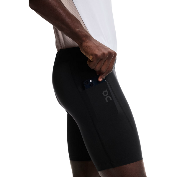 En person iført On 8 Performance Tights, Herre, Black lægger en smartphone i en sidelomme. Strømpebukserne vises på en almindelig hvid baggrund, hvilket understreger deres slanke og funktionelle design med det synlige ON-logo.