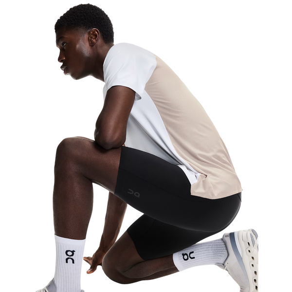 En person i en beige og hvid sportstrøje, sorte shorts over On 8 Performance Tights til mænd i sort, hvide sokker og hvide sneakers knæler på et knæ mod en almindelig hvid baggrund.