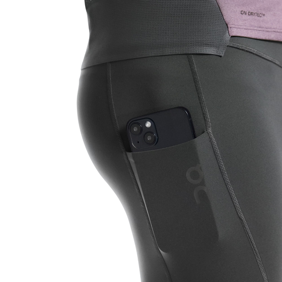Nærbillede af en person iført On 8" Performance Tights, Herre, Eclipse med en smartphone i sidelommen; en del af linningen på en lilla top er også synlig.