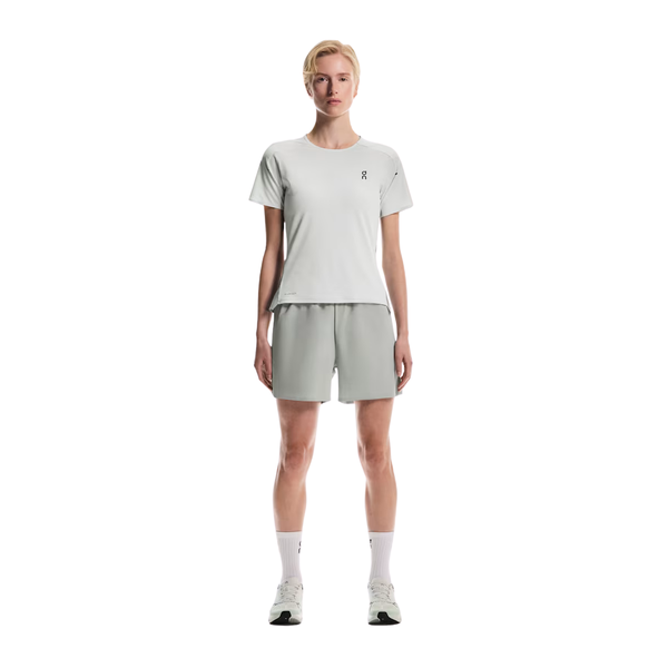En person står med ansigtet fremad iført en On Performance-T Dame i Iceberg/Tin, matchende shorts, hvide strømper og hvide sneakers. Den almindelige hvide baggrund fremhæver den atletiske On DryTec-stil.