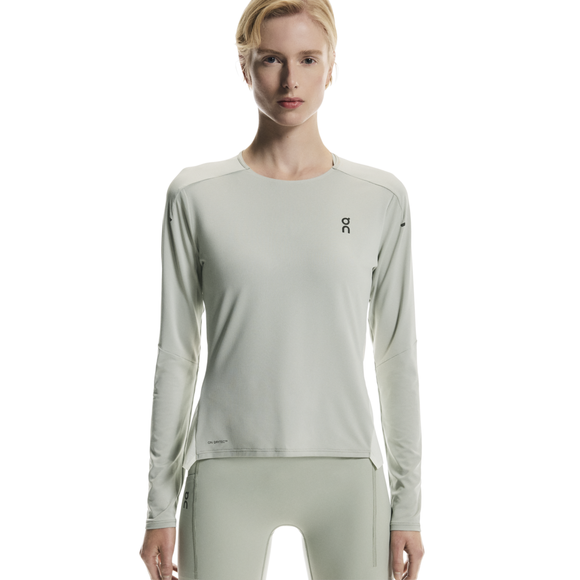 En kvinde med kort blondt hår bærer On Performance Long-T til kvinder i Tin/Iceberg og matchende leggings. Den lysegrå, svedtransporterende top har et lille On-logo på venstre bryst og højre skulder. Baggrunden er almindelig hvid.