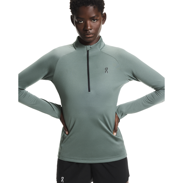 En kvinde står med hænderne på hofterne, iført On Climate Shirt, Dame, Olive og sorte shorts. Baggrunden er helt hvid.