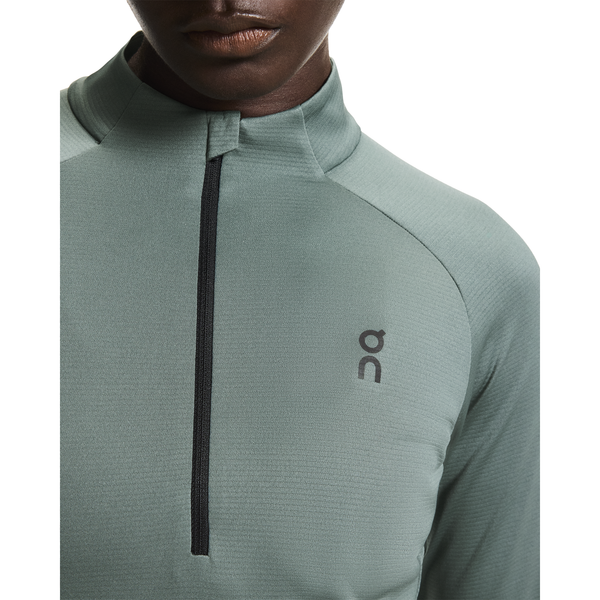 Nærbillede af en person i On Climate Shirt, Dame, Olive - en lysegrøn fugttransporterende løbetrøje med sort kvart lynlås og On-logo på venstre bryst - delvist vist mod en hvid baggrund.