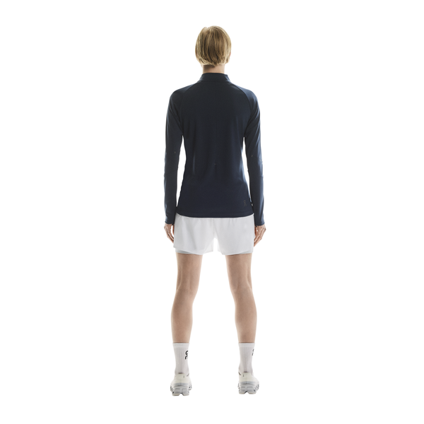 En kvinde står med ryggen til kameraet iført On Climate Shirt, Dame, Navy, stylet med hvide shorts, sokker og sneakers mod en almindelig hvid baggrund.