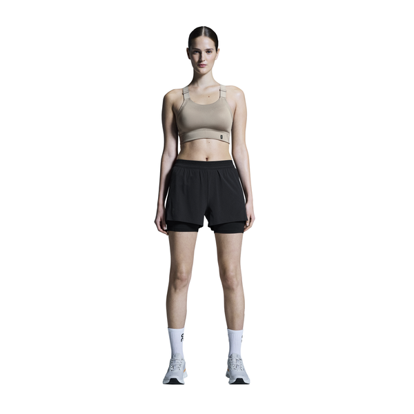En person står med ansigtet fremad i beige træningstøj - en sports-bh og On 3" Performance 2/1 Shorts til kvinder i sort - sammen med hvide sokker og lyse sneakers på en almindelig hvid baggrund.