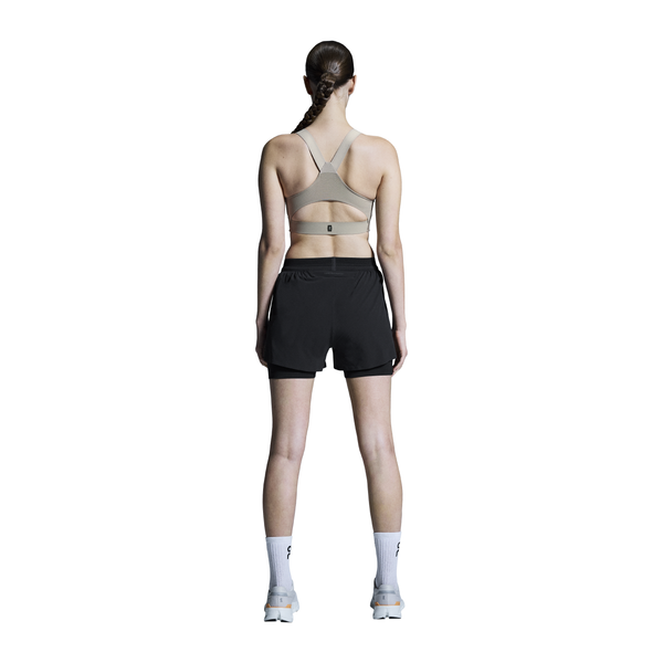 En person med langt tilbagestrøget hår står med ansigtet vendt mod en hvid baggrund iført On 3" Performance 2/1 Shorts, Dame, Black - ideal træningstøj for løb dame - sammen med en sports-bh, hvide sokker og sportssko.