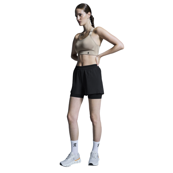 En kvinde står på en hvid baggrund i træningstøj: beige sports-bh, On 3" Performance 2/1 Shorts Dame Black, hvide strømper og sneakers. Hendes hår er flettet, og hendes hænder hviler på lænden - perfekt til enhver løb dame.