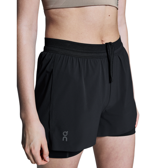 En person iført en beige sports-bh og On 3" Performance 2/1 Shorts, Dame, Black med On-logoet på venstre ben står på en hvid baggrund og viser smart træningstøj til løb dame. Kun overkroppen og det øverste af benene er synlige.