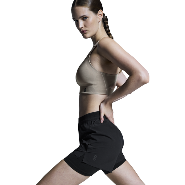 En kvinde med en lang fletning bærer sorte On 3" Performance 2/1 Shorts til kvinder og en sports-bh, mens hun står i sideprofil med en hånd på hoften mod en almindelig hvid baggrund.