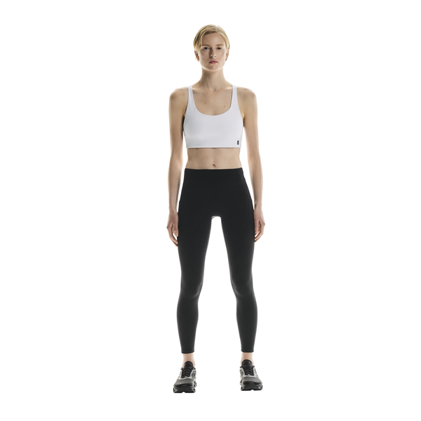 En person står med front mod en almindelig hvid baggrund iført en hvid sports-bh, sorte On Performance Tights Winter Dame og sorte sportssko.
