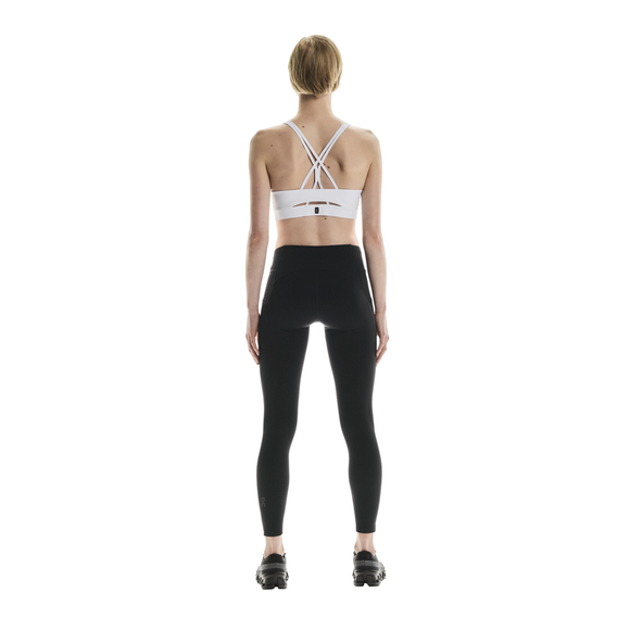 En person står med ansigtet vendt mod kameraet iført en hvid sports-bh med stropper, sorte On Performance Tights Winter til kvinder og sorte sportssko på en almindelig hvid baggrund.