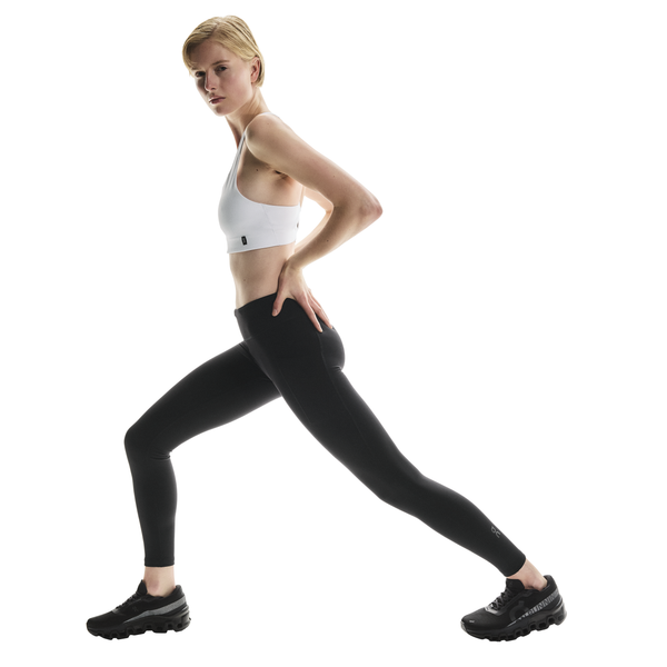 En person med kort blondt hår iført hvid sports-bh, On Performance Tights Winter, Dame, Black og sorte sportssko udfører et forlæns udfald mod en almindelig hvid baggrund.