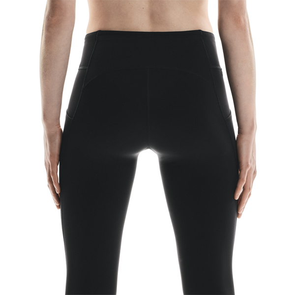 En person ses bagfra iført On Performance Tights Winter, Dame, Black, med synlige sømme og sidelommer. Armene hviler i siden mod en almindelig hvid baggrund, som fremhæver det slanke design.